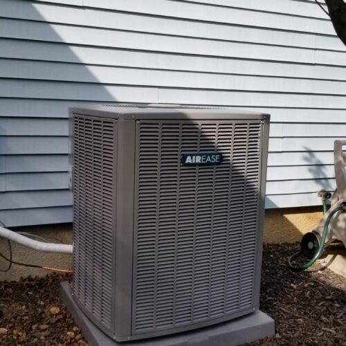 A gray AirEase outdoor air conditioning unit sits on a concrete base next to a house with light blue siding. The surrounding area has mulch, a green hose, and some plant leaves overhead.