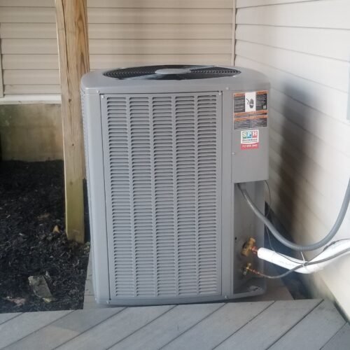 A gray outdoor air conditioning unit is installed next to a house on a wooden deck, with a wooden post and siding visible nearby.