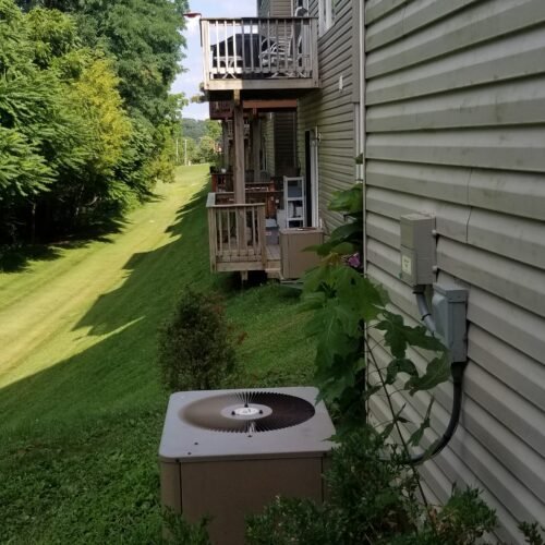 A row of apartment balconies extends along the side of a beige building with siding. An outdoor air conditioning unit sits amidst green bushes, and a grassy area slopes gently beside the building, bordered by trees.