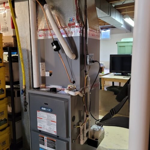 A gray Airease furnace unit installed in a basement, surrounded by pipes, ducts, insulation, and storage bins. A TV and green storage tub are visible in the background.