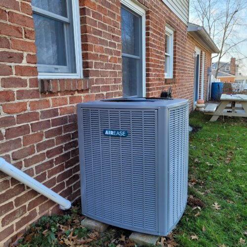 A gray AirEase outdoor HVAC unit sits on a concrete pad next to the brick exterior wall of a house, with windows above and fallen leaves scattered on the grass nearby.