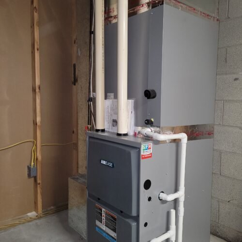 A gray furnace unit with white PVC pipes is installed in a basement next to a concrete wall and exposed wood framing. Yellow electrical wires and a gray electrical outlet are visible on the unfinished wall.