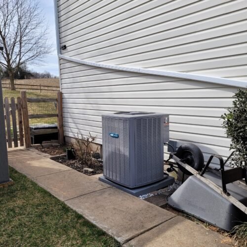 A gray outdoor air conditioning unit sits on a concrete slab next to a white house. A tipped-over wheelbarrow and shrub are nearby, with a small garden bed and grass in the background.