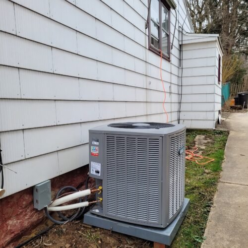 A gray outdoor air conditioning unit is installed next to a white house with horizontal siding. Electrical cables and pipes connect the unit to the house. The unit sits on a concrete pad beside a sidewalk.