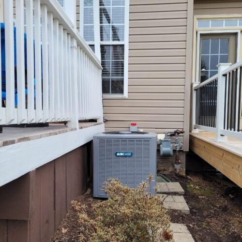 A gray AirEase HVAC unit is installed between a house with tan siding and a wooden porch. Concrete pavers lead to the unit, and a small bush is visible in the foreground.