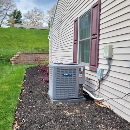 A gray AirEase air conditioning unit is installed next to a beige house with maroon shutters. The unit sits on mulch beside a landscaped yard with grass, plants, and a stone retaining wall in the background.