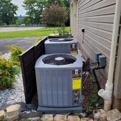 Two outdoor air conditioning units sit next to a house with siding, separated by a black privacy screen, amid landscaping with rocks, bricks, and bushes under a partly cloudy sky.