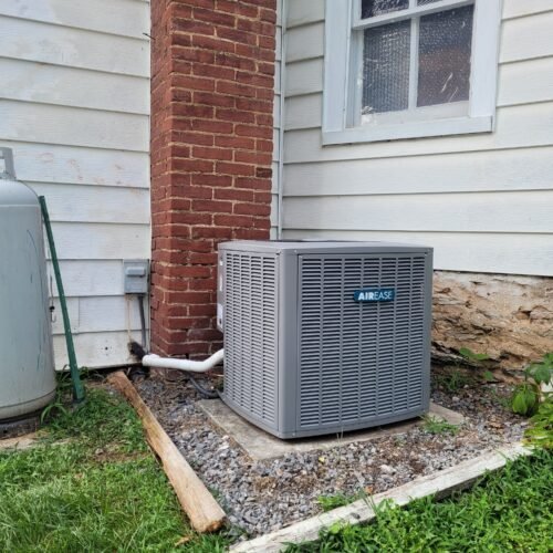 A gray AIR-EASE air conditioning unit sits on a gravel bed next to a brick chimney and white-sided house with a curved window. Green grass and a propane tank are visible nearby.