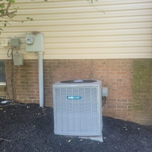 An outdoor Airease air conditioning unit sits on a concrete pad next to a brick and siding house. Nearby, utility boxes and a power meter are mounted on the wall above dark mulch and greenery.