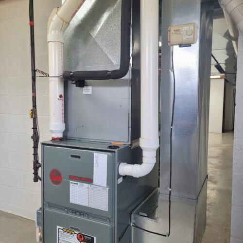 A Rheem gas furnace with white PVC vent pipes is installed in a basement with concrete floors and unfinished walls. The furnace has warning labels and an attached thermostat on the ductwork.