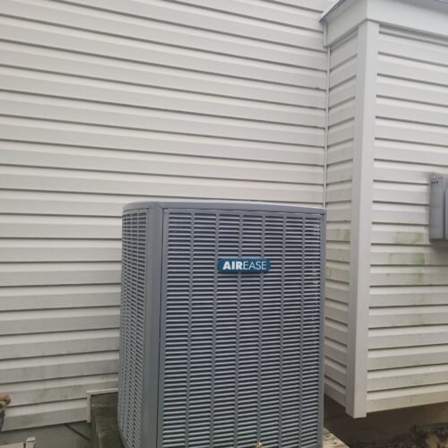 A gray Airease air conditioning unit is installed on a small platform next to the white vinyl-sided exterior wall of a house, with some green plants in the foreground.