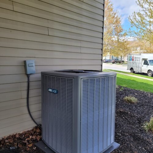 A gray outdoor AirEase air conditioning unit sits on a concrete pad next to a beige-sided house, with mulch and soil bags nearby. Green grass, trees, and a white van are visible in the background.