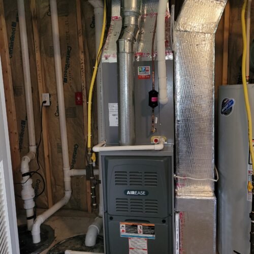 A residential furnace unit labeled "AirEase" is installed in a basement utility room with exposed insulation, ductwork, yellow electrical wires, and a nearby water heater. Various pipes and vents connect to the furnace.