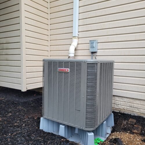 A gray Lennox air conditioning unit is installed outside a beige house, sitting on a plastic platform with a drain pipe above it, surrounded by mulch and some green marking on the ground.