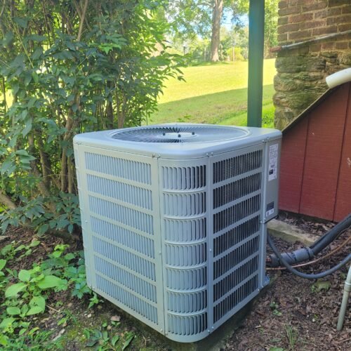An outdoor air conditioning unit sits on the ground next to a house, surrounded by green plants and grass, with a mowed lawn and trees visible in the background.