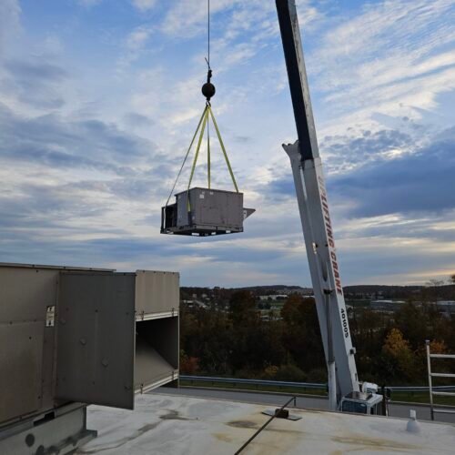 A crane lifts a large HVAC unit onto a rooftop. The unit is secured with yellow straps, and the sky is partly cloudy with a view of trees and buildings in the background.