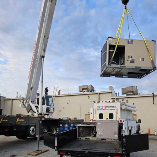 A large crane lifts an industrial HVAC unit onto a flatbed truck beside a commercial building, while workers oversee the operation on a cloudy day.