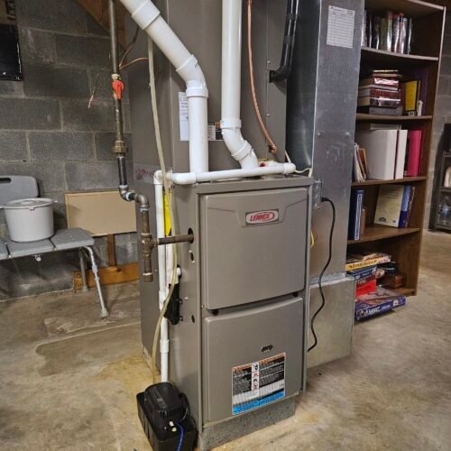A Lennox furnace in a basement, surrounded by pipes and ducts. Nearby are a gray folding chair and a wooden shelf filled with books, board games, and other items. The walls are unfinished concrete blocks.