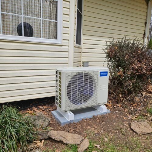 An outdoor air conditioning unit is installed on a concrete pad next to a beige house with siding, near a window with curtains and surrounded by bushes, grass, and fallen leaves.