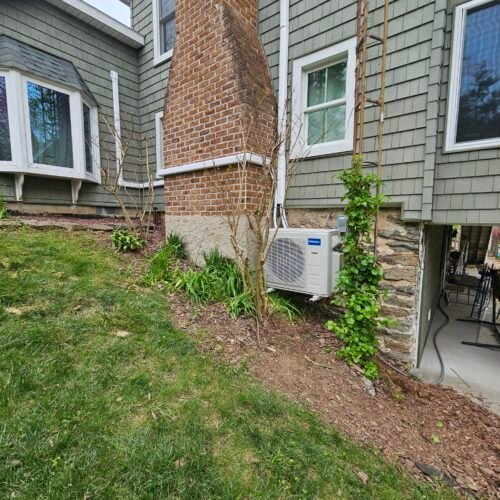 A heat pump outdoor unit is mounted on the side of a green house with brick chimney. The yard has grass, mulch, plants, and a small patio area is visible under the house.