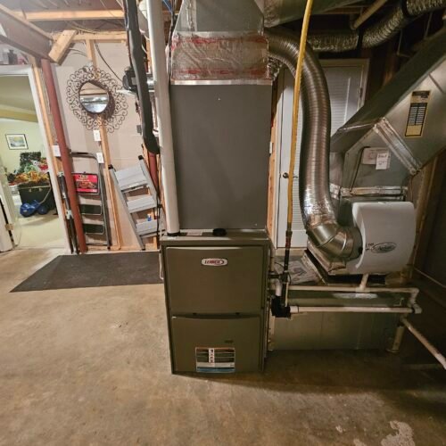 A central heating furnace unit with ductwork is installed in a basement. The basement has concrete floors, an open doorway, and a decorative wall mirror is visible in an adjacent room.