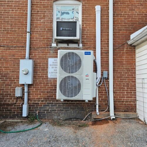 An air conditioning unit is mounted on a brick wall beneath a window. There are pipes, an electrical box, and a hose nearby. A sign on the wall reads, "NO SKATEBOARDING OR BIKE RIDING, NO LOITERING.