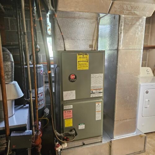A Rheem HVAC unit is installed in a basement next to a water heater and a washing machine. Metal ductwork and various pipes surround the area. The walls are partially concrete blocks. Sunlight comes from a small window.