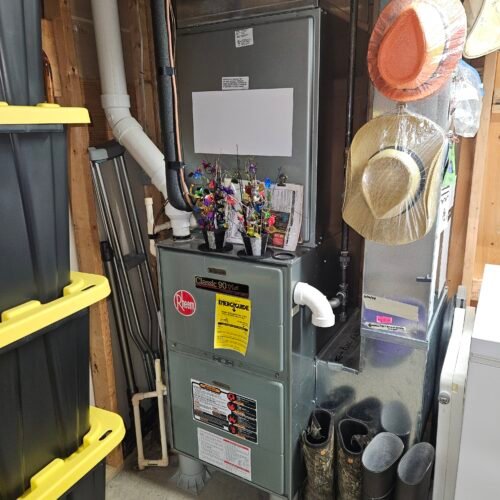 A furnace unit in a utility room with hats hanging on the side, storage bins stacked nearby, boots on the floor, and artificial flower decorations placed on top of the furnace.