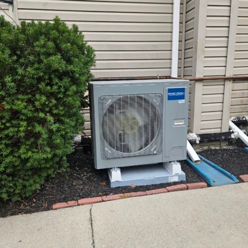 A gray MRCOOL air conditioning unit is installed outside next to a beige building, partially surrounded by green bushes and landscaping with a sidewalk in front.