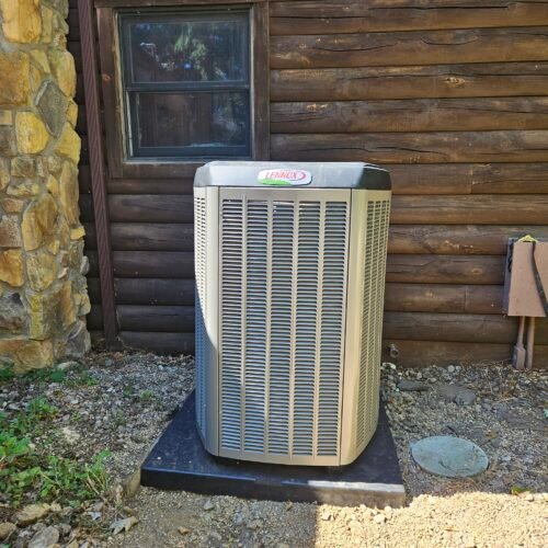 A Lennox air conditioning unit sits on a concrete pad outside a wooden log cabin, with a stone wall and a window visible in the background.