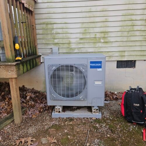 An outdoor HVAC unit labeled “MRCOOL” sits on concrete blocks next to a house with a stained siding wall. Nearby are a red tool bag and a stair railing with a yellow and black drill resting on it. Fallen leaves cover the ground.