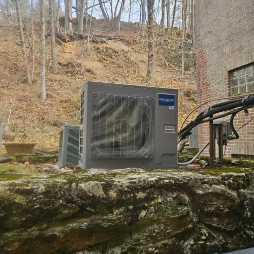 An outdoor heat pump unit sits on a stone platform next to a brick building, with trees and a steep, leaf-covered hillside in the background. Several cables and pipes are connected to the unit.