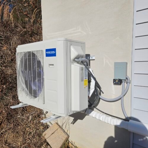 A white MrCool outdoor air conditioning unit is mounted on a wall, with electrical and refrigerant lines connected and running into a gray junction box; dry grass and sunlight are visible in the background.