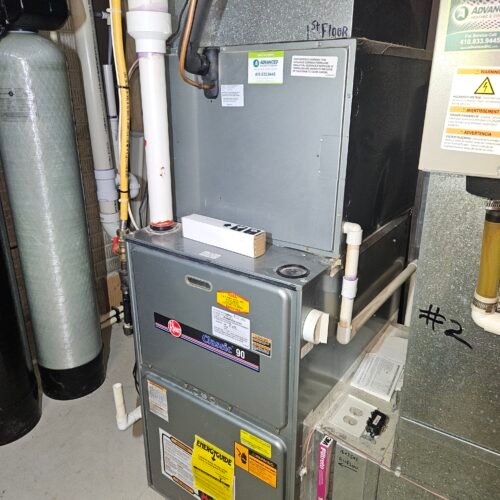 A Rheem Classic 90 gas furnace installed in a utility room, surrounded by pipes, ductwork, and a nearby water softener system. A small white box sits on top of the furnace unit.