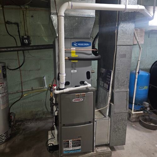 A gray Lennox furnace with white PVC pipes installed in a basement, connected to ductwork and surrounded by other equipment including a water heater and a blue pressure tank. The walls are painted green.
