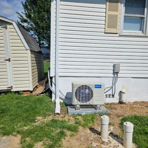An outdoor air conditioning unit is installed next to a white house with beige shutters, near three white capped pipes and a shed on a grassy yard under a clear, sunny sky.