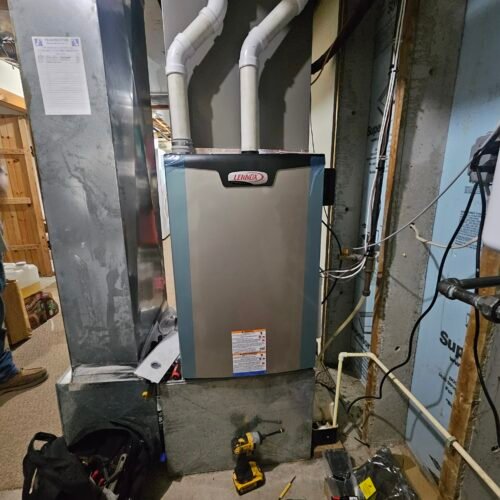A Lennox furnace installed in a basement, surrounded by pipes and tools. The unit sits on a concrete base, with ducts and wiring visible nearby, and a toolkit and power drill are on the floor in front.