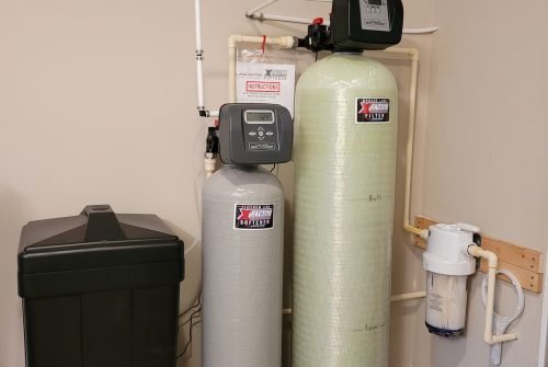 A water treatment system setup with two tall cylindrical tanks, a black brine tank, and a white wall-mounted filter connected by pipes, installed in a room with beige walls and tile flooring. A water treatment system setup with two tall cylindrical tanks, a black brine tank, and a white wall-mounted filter connected by pipes, installed in a room with beige walls and tile flooring.
