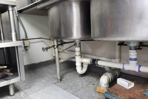 Two large metal sink basins with visible plumbing pipes underneath, connected by white PVC pipes. The area has gray tiled flooring, an electrical outlet on the wall, and a small white box on the floor. Two large metal sink basins with visible plumbing pipes underneath, connected by white PVC pipes. The area has gray tiled flooring, an electrical outlet on the wall, and a small white box on the floor.