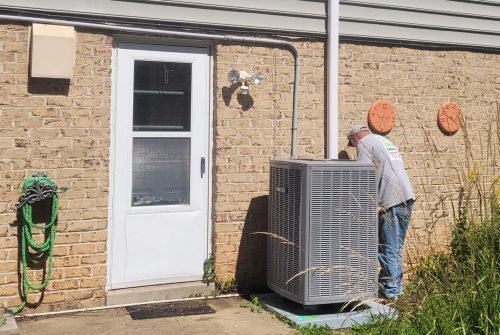 A person wearing jeans and a t-shirt repairs or inspects a large outdoor HVAC unit next to a brick and siding house on a sunny day, with green grass and some overgrown plants nearby. A person wearing jeans and a t-shirt repairs or inspects a large outdoor HVAC unit next to a brick and siding house on a sunny day, with green grass and some overgrown plants nearby.