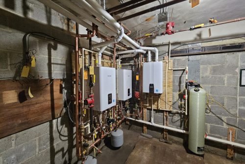 A basement utility room with exposed pipes, two mounted white boilers, water tanks, a filtration system, control valves, and cinder block walls under bright overhead lighting. A basement utility room with exposed pipes, two mounted white boilers, water tanks, a filtration system, control valves, and cinder block walls under bright overhead lighting.