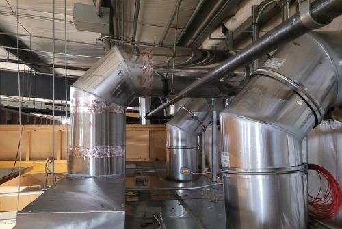 Large silver metal air ducts and pipes run along the ceiling in an industrial building, resembling robust residential HVAC systems. The ducts are joined with red tape, surrounded by electrical conduits and yellow insulation, giving an unfinished appearance. Large silver metal air ducts and pipes run along the ceiling in an industrial building, resembling robust residential HVAC systems. The ducts are joined with red tape, surrounded by electrical conduits and yellow insulation, giving an unfinished appearance.